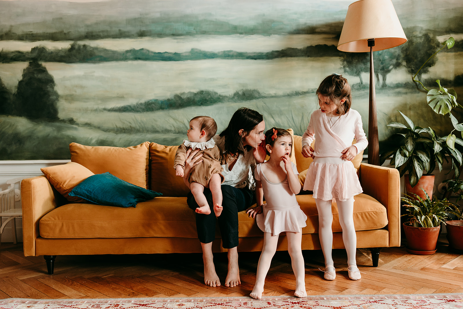 maman sur canapé jaune, parle à ses filles en tutu et a son bébé dans les bras